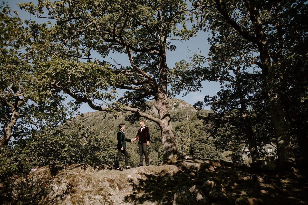 Two grooms pictured beneath the trees at lakeside venue